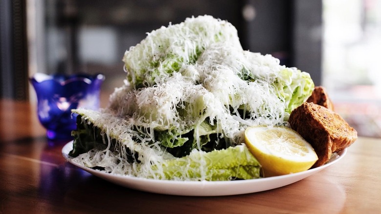 A Caesar salad draped in shredded Reggianito with half of a lemon and large croutons on a wooden table