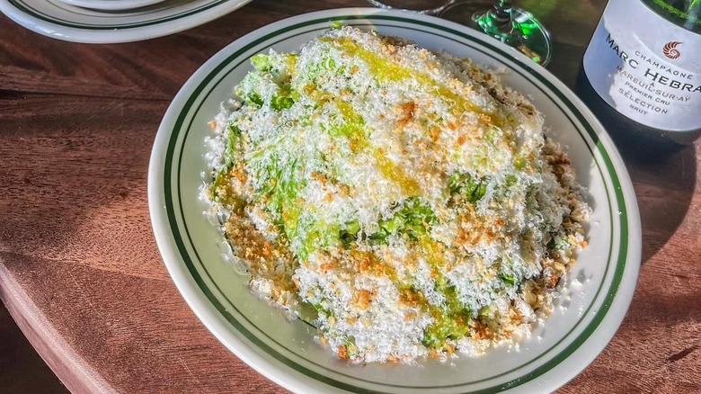 A Caesar salad in a white bowl on a wooden table next to a bottle of Champagne