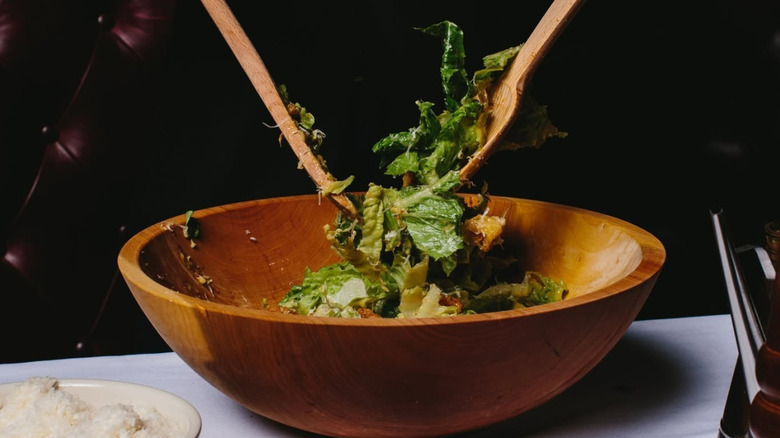 A Caesar salad being made tableside with wooden spoons in a wooden bowl