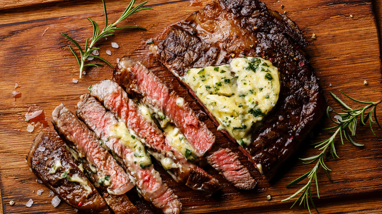 An overhead short of a ribeye steak placed on a wooden cutting board, decorated with herby butter
