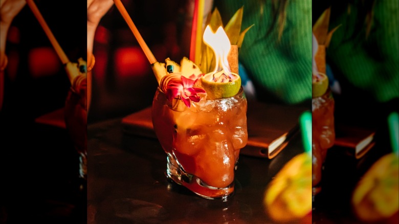 Two people drinking from a red skull-shaped drink with garnishes and a flaming lime half