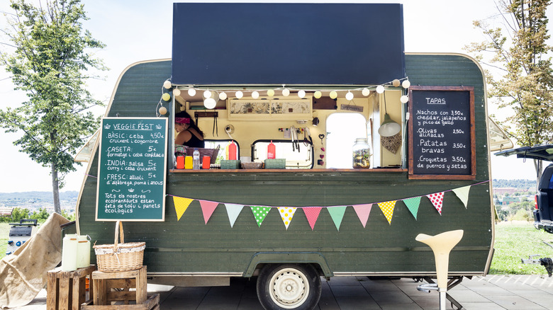 A grey food truck in a park with flags and string lights decorating it