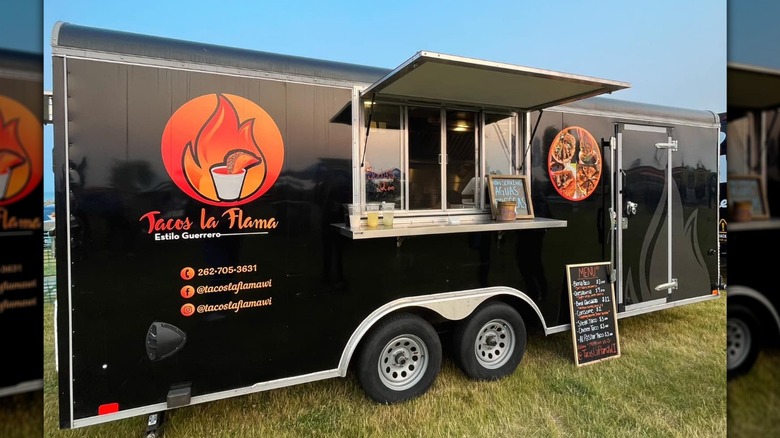 The black Tacos La Flama food truck with a menu board outside
