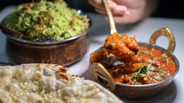 Spoon dipping into a bowl of butter chicken with dishes of naan and biryani nearby