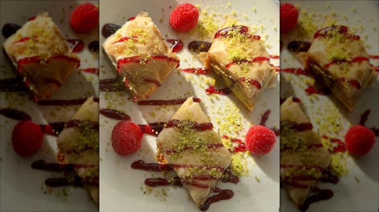 Three pieces of baklava drizzled with raspberry sauce, garnished with fresh raspberries and ground pistachios on a white plate at Blue Olive Mediterranean Cuisine