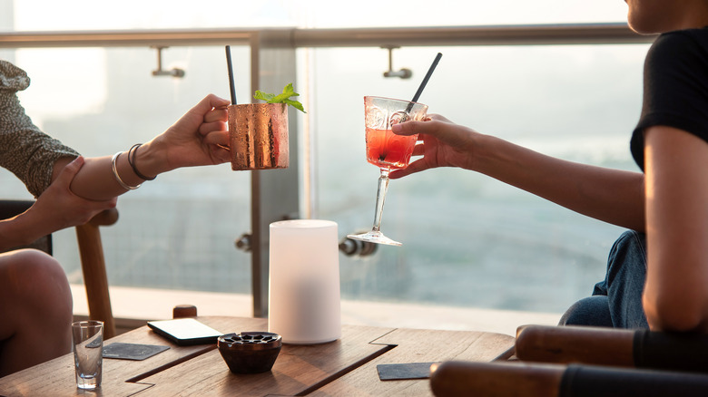 Two women sitting at a rooftop bar about to clink their drinks together
