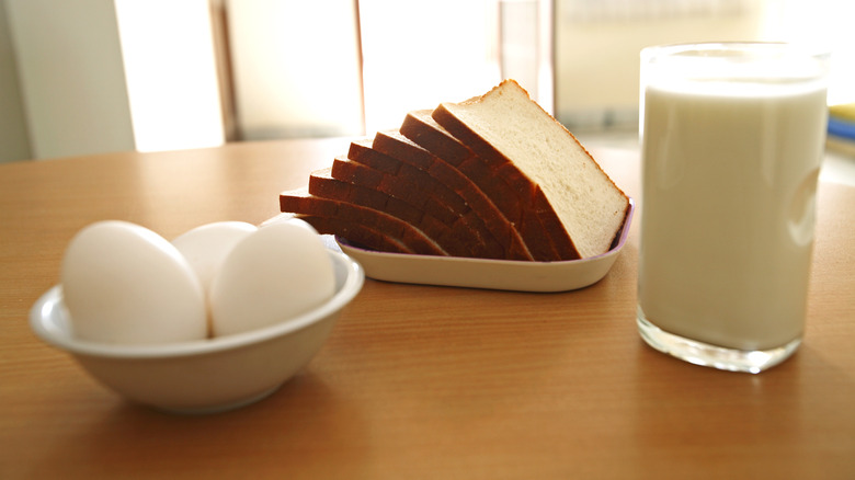 A bowl with three eggs, a plate of sliced bread, and a glass of milk sit on top a wooden table