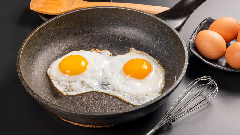 Two fried eggs in a nonstick pan with whisk and whole eggs beside it.