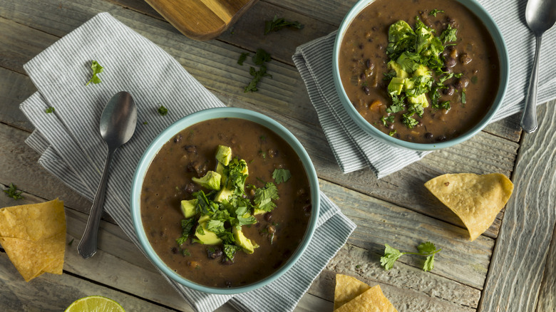 Two blue bowls of black bean soup garnished with avocado and cilantro on napkins next to spoons and tortilla chips