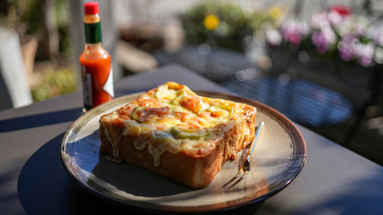 A piece of pizza toast on a plate with two-pronged fork and a bottle of hot sauce in the background