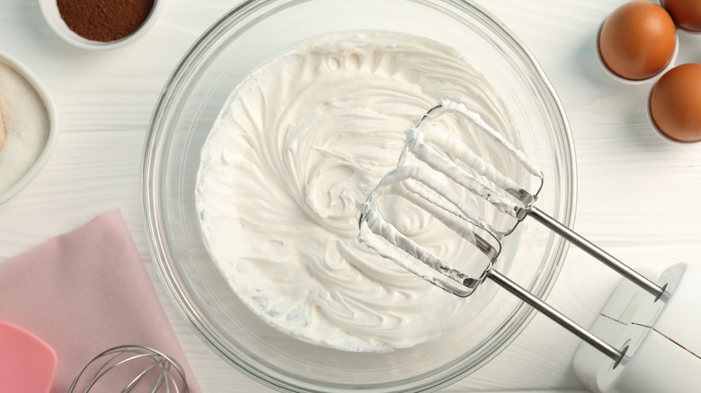 A bowl of freshly whipped cream with the mixer used just above the bowl and eggs and spices seen on the table