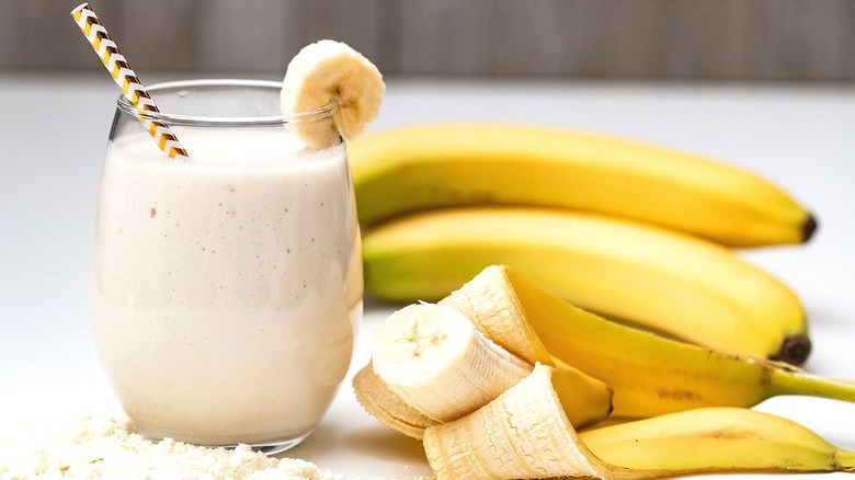 Banana milkshake with a gold and white straw in a short clear glass next to peeled banana
