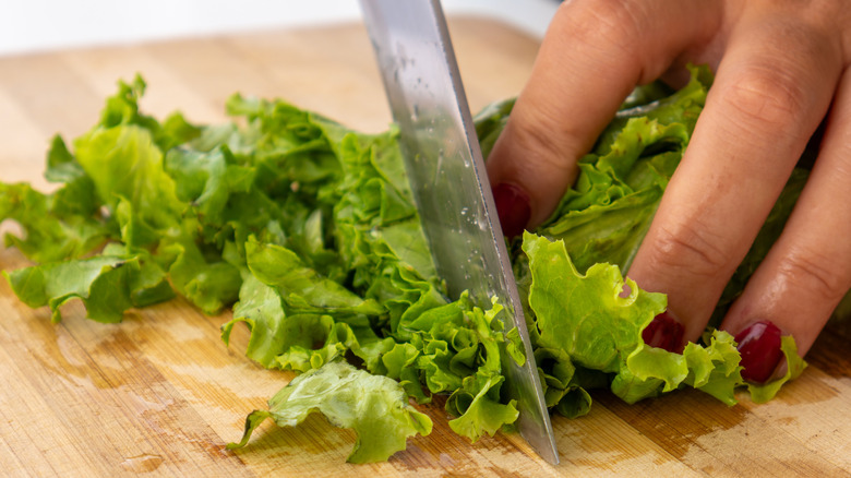 A hand using a chef's knife to chop fresh lettuce on a cutting board.