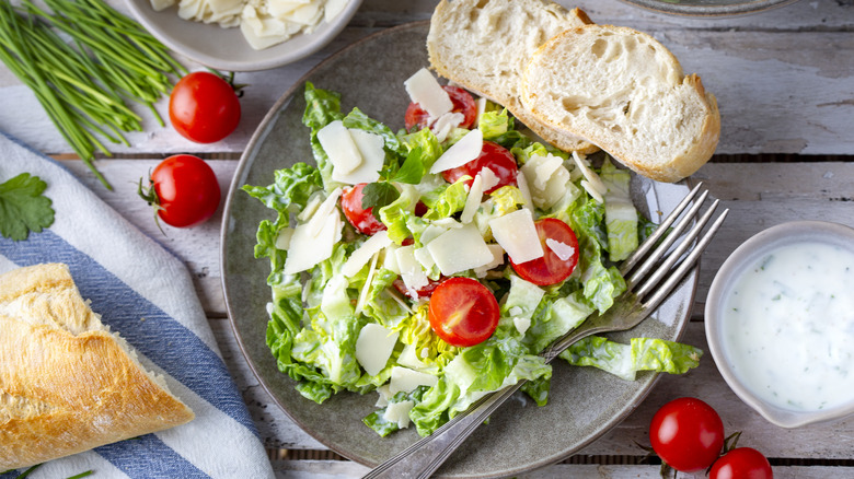 A chopped romaine salad with cherry tomatoes and shaved parmesan in a bowl with sliced bread, surrounded by salad dressing, tomatoes, and herbs.