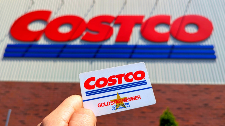 A person's hand holding a Costco gold star member card in front of a costco store sign