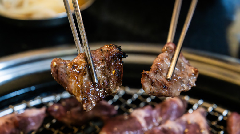 Two pairs of stainless steel chopsticks holding slices of charred meat above a grill