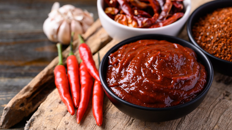 A bowl of gochujang on a wooden board surrounded by chili peppers, garlic, and chili powder