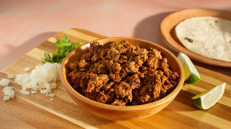 A bowl of taco meat arranged with chopped onion, cilantro, and lime wedges on a cutting board.