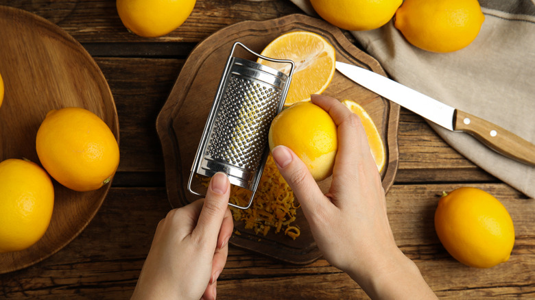 Close up of hands zesting a lemon over small wood plate with other whole lemons in the background