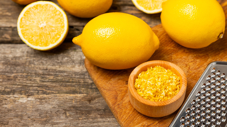 Small wood bowl of lemon zest next to whole lemons and grater on cutting board