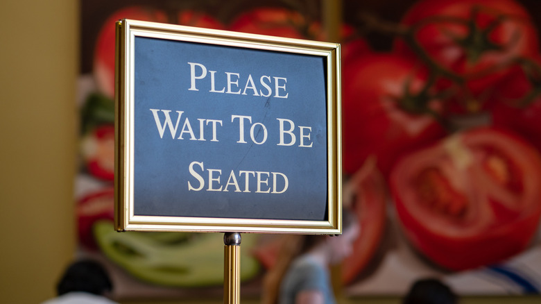 A black and gold 'please wait to be seated' sign in a restaurant