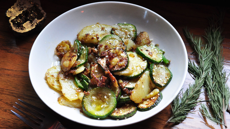 White shallow bowl of roasted caramelized zucchini slices with a few sprigs of rosemary