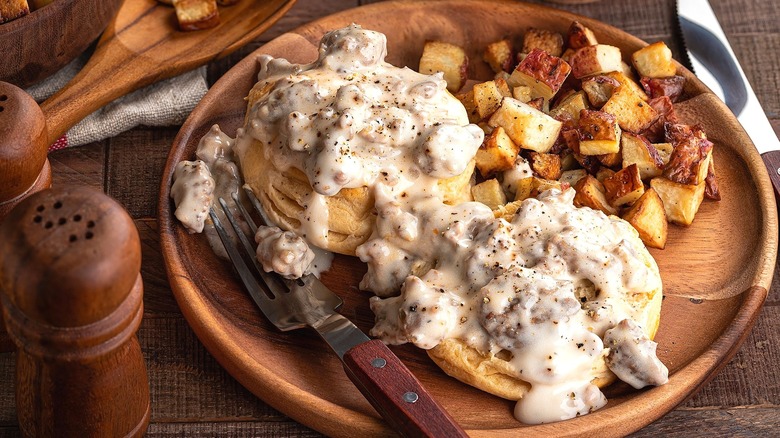 Two biscuits covered in sausage gravy and roasted potatoes on a wooden plate with a fork