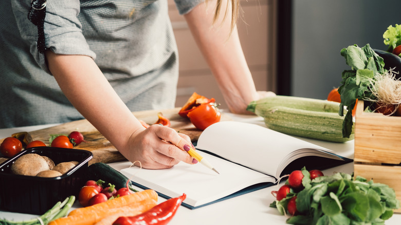 Person reading and editing a recipe in a notebook at a kitchen counter covered in fresh produce