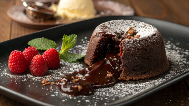 A freshly cut, molten chocolate cake, the lava flowing out, on a black plate with raspberries and a sprig of time, vanilla ice cream in the background.