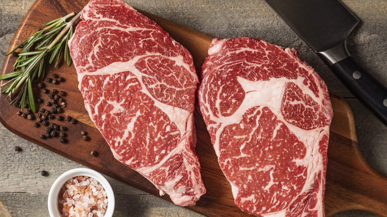 Two raw wagyu steaks on a cutting board surrounded by herbs and seasonings