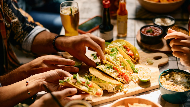 Hands digging into a platter of tacos with beer and hot sauce in the background