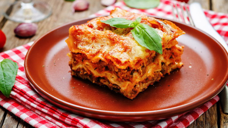 A slice of lasagna with basil shown on a folded red tablecloth on a picnic table