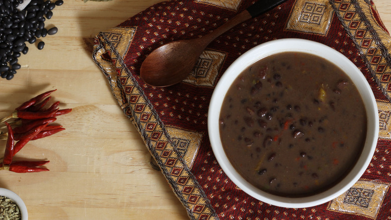 A white bowl of black bean soup on dark decorative placemat with wood spoon.