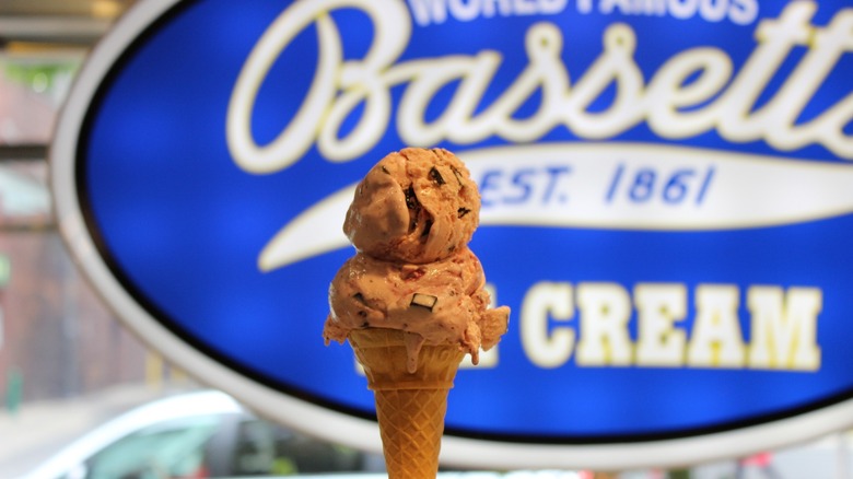 Person holding a double-scoop cone in front of Bassetts sign
