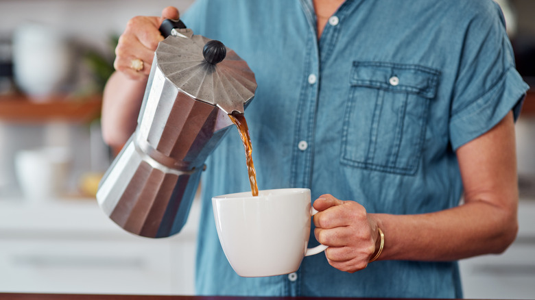 A woman pours coffee from a Moka pot into a cup.