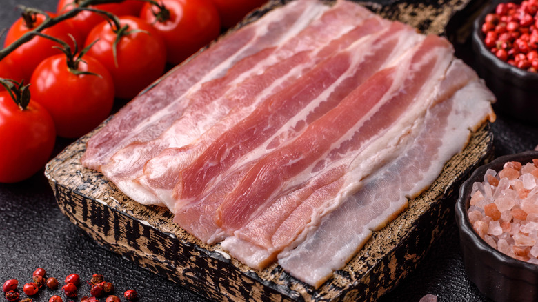Slices of raw bacon on a cutting board with tomatoes, peppercorns, and salt in the background