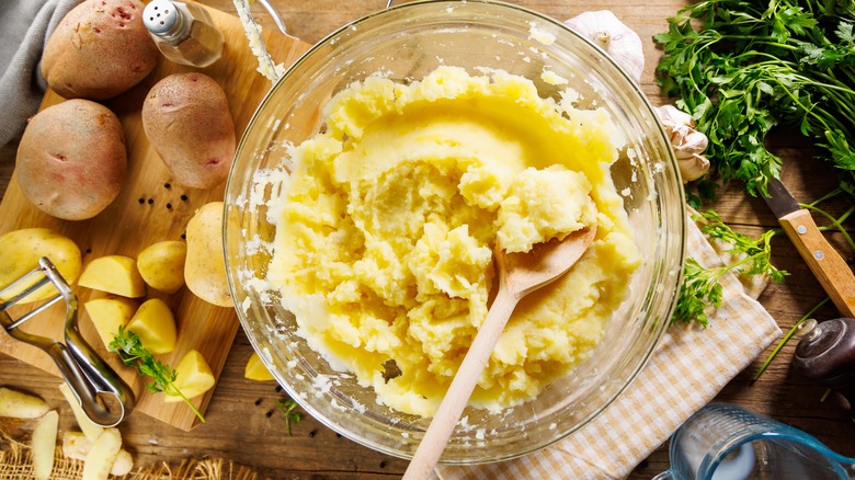 Mashed potatoes in a bowl with a wooden spoon with potatoes and fresh herbs on the side