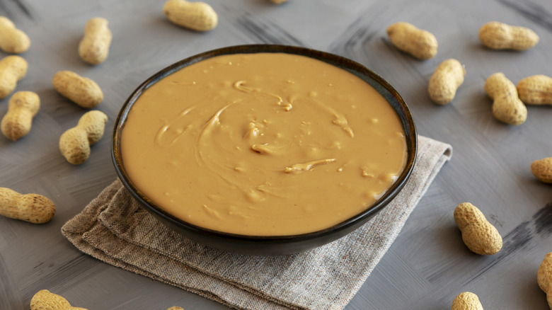 Small dark bowl of natural peanut butter sitting on folded cloth with whole peanuts around it