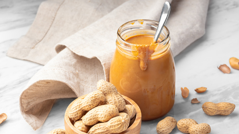 Open glass jar of natural peanut butter spilling over the rim with a spoon and whole peanuts in small wood bowl next to a beige cloth.