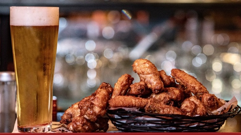 A beer served in a tall glass next to a basket of chicken hot wings on a bar top.