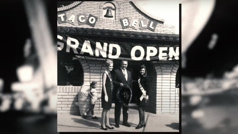 Black-and-white photo of Glen Bell and two women in pageant sashes in front of the first Taco Bell location with its grand opening banner