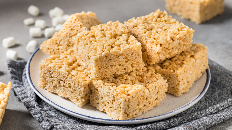 A plate with several Rice Krispies Treats sitting on a table with unused marshmallows visible in the background