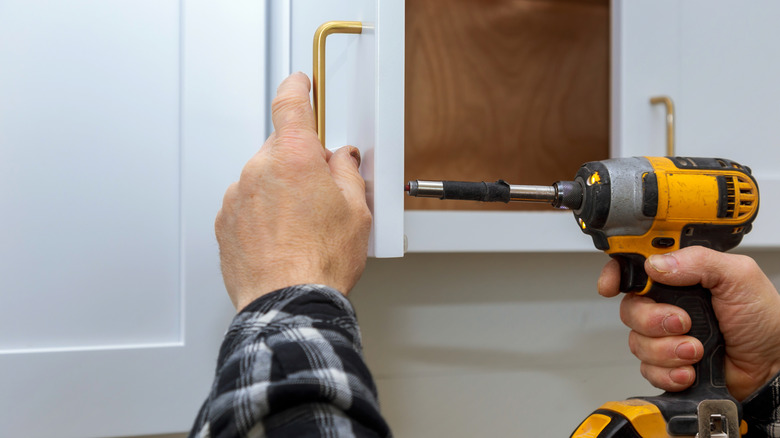 Hands and arms of person in plaid shirt changing handles on white cabinets with a drill