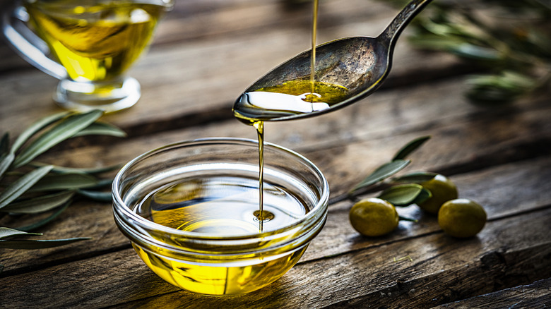 Extra virgin olive oil is poured into a spoon over a glass bowl on a rustic wooden table.