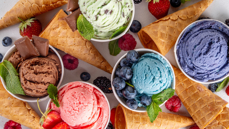 Small bowls of ice cream in an array of colors and flavors with relating garnishes — chocolate, strawberry, mint chocolate chip, blueberry, and more, surrounded by waffle cones and fruits.