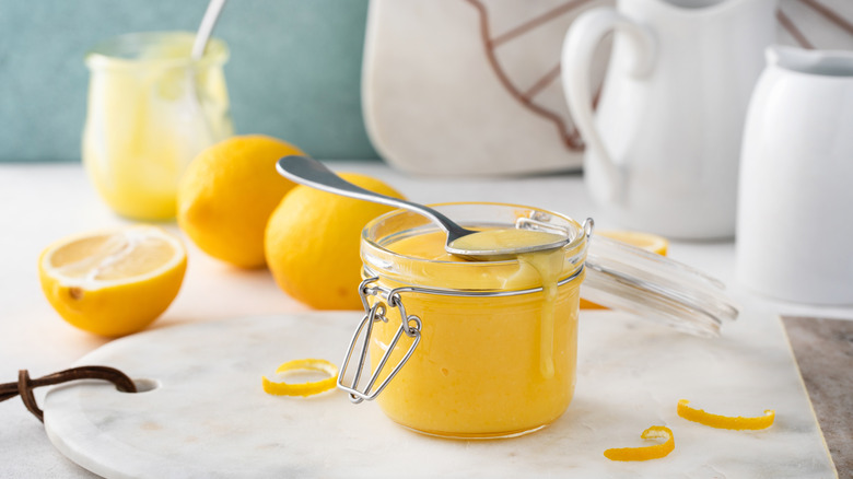 A jar of fresh lemon curd on a marble cutting board with lemons in the background.
