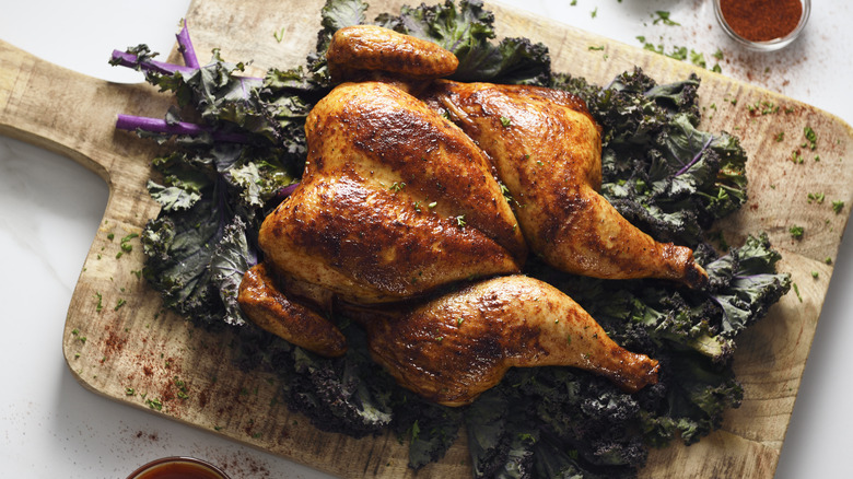A perfectly roasted whole chicken on a bed of kale over a wooden cutting board, small bowls of spices and herbs in the background.