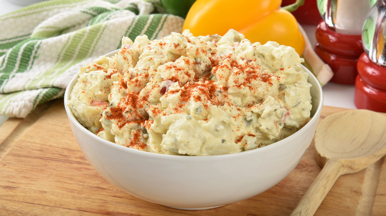 A bowl of mayo and mustard potato salad, topped with paprika on a wooden cutting board, a wooden spoon to the side, a tea towel in the background along with bell peppers.