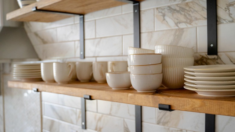 Open wood and metal shelving in a kitchen stacked full of white and beige bowls, plates, and mugs