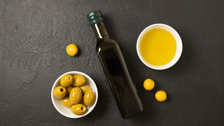 A bottle of olive oil next to olive oil and green olives in a white bowl